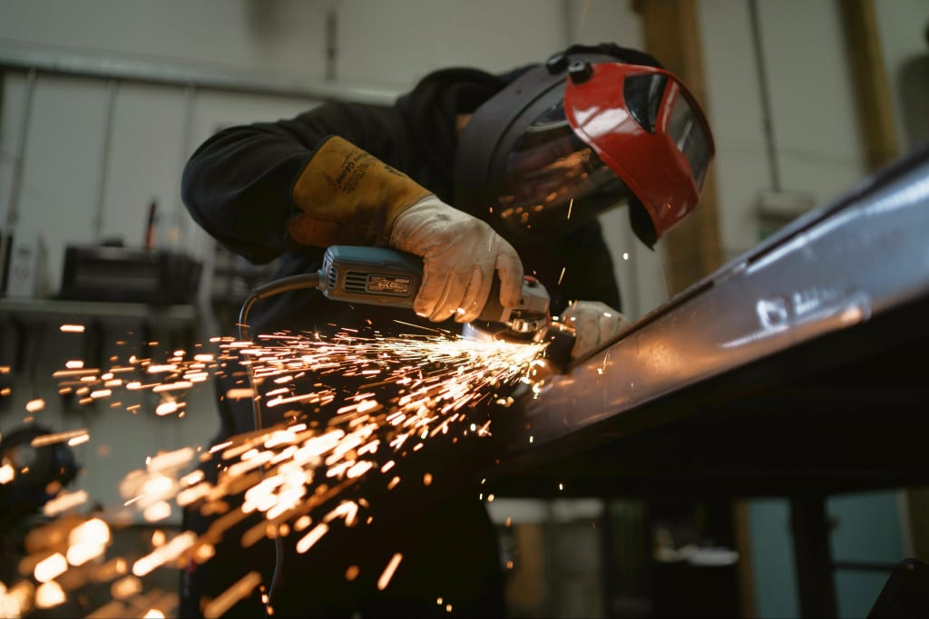 A worker in a welding mask grinding a steel beam, throwing sparks across the workshop floor